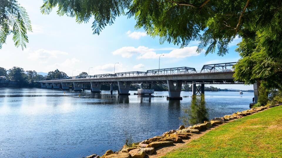 A bridge over a calm river with a grassy bank and trees in the foreground.