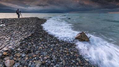 A rocky shoreline with a person standing on the left, a tripod, and a tripod with a camera on it.