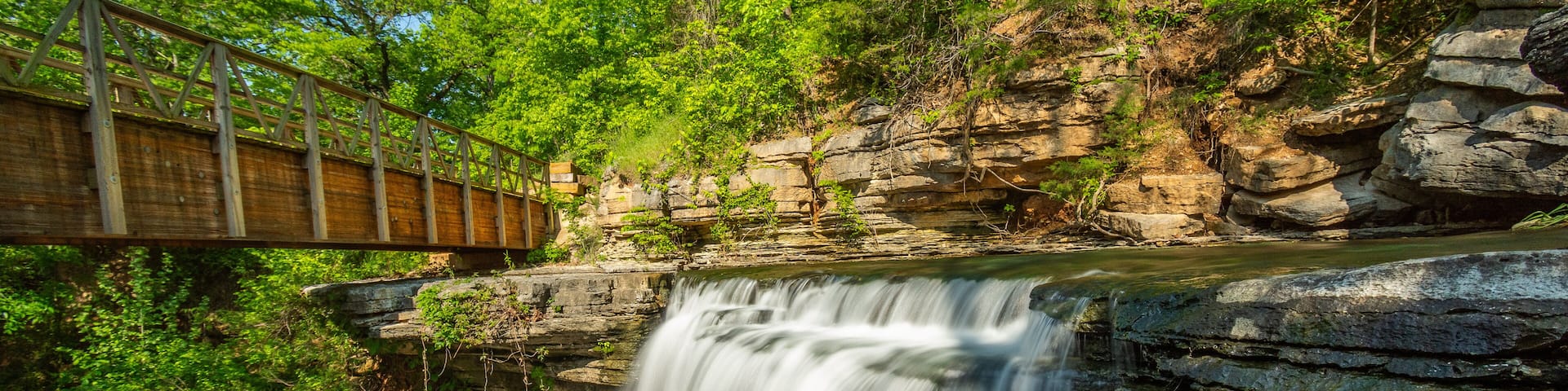A wooden bridge over a waterfall with surrounding greenery and rocky cliffs.