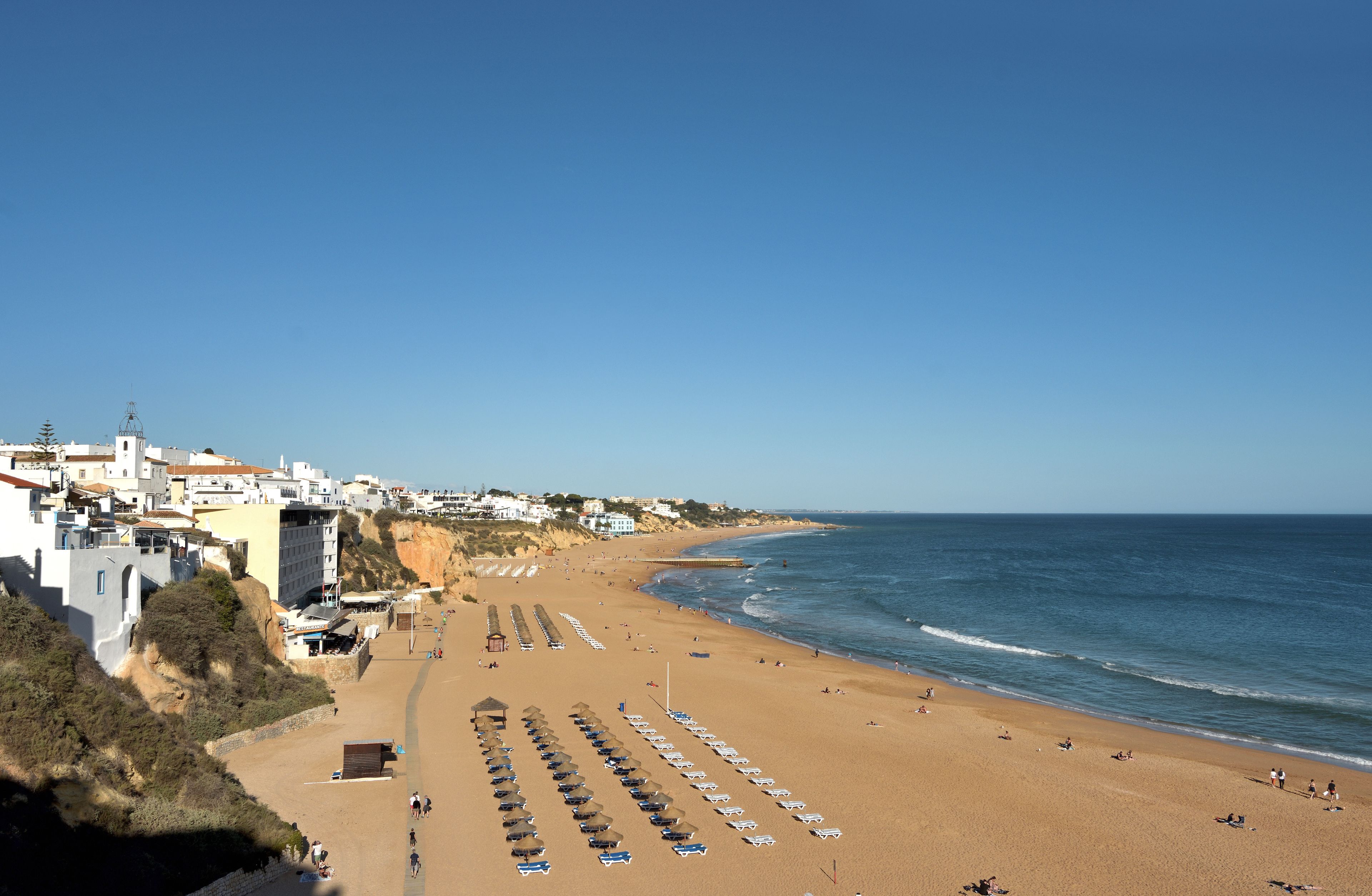 Un pueblo costero con una playa arenosa, hileras de sombrillas y un cielo azul despejado.