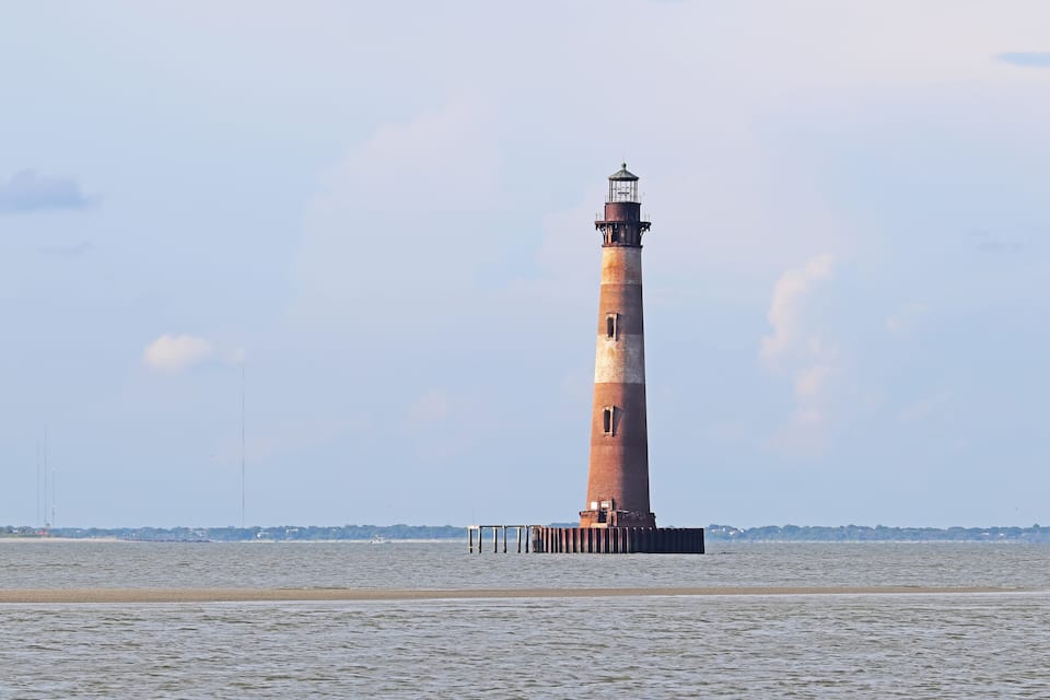 Um farol alto e esguio com padrão listrado em vermelho e branco, erguido sobre um cais em uma massa de água.