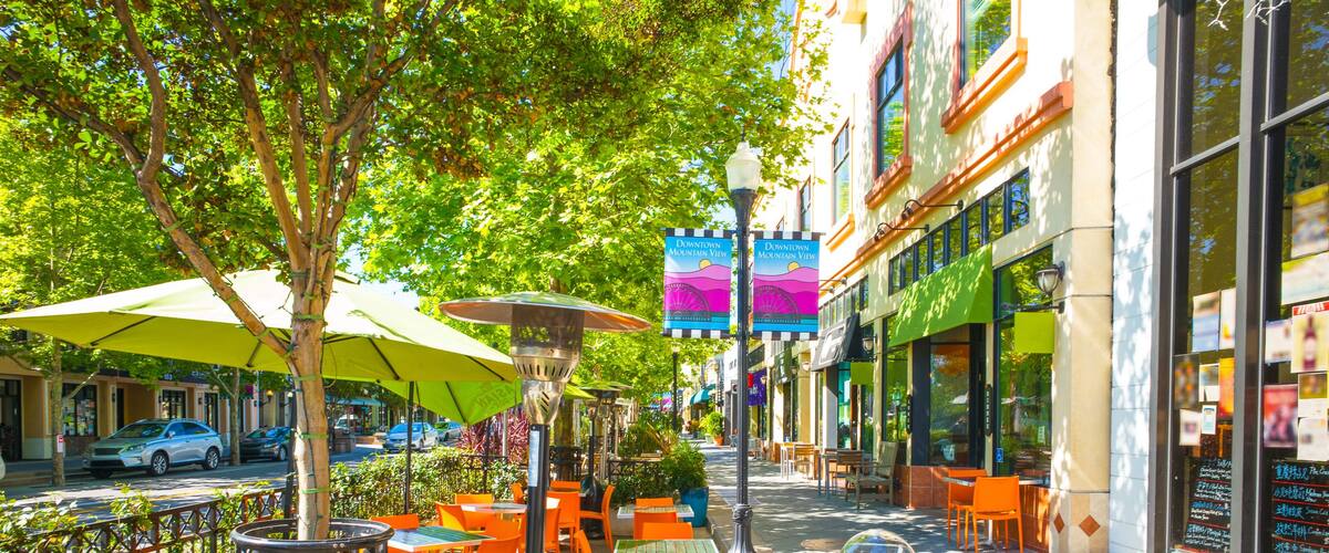 An outdoor cafe with orange tables and green umbrellas, surrounded by trees and buildings.