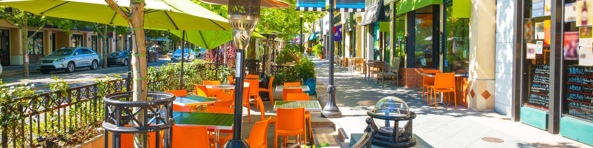 An outdoor cafe with orange tables and green umbrellas, surrounded by trees and buildings.