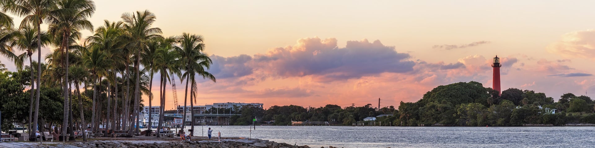 A sunset over a beach with palm trees, a lighthouse, and a rocky shoreline.