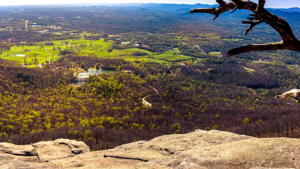A mountainous landscape with a clear view of a valley, a winding road, and a large rock formation in the foreground.
