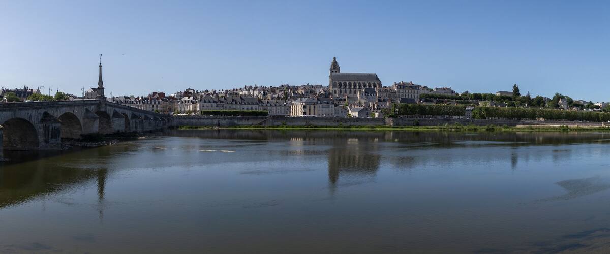 A river with a stone bridge, a large domed building, and a densely packed cityscape in the background.