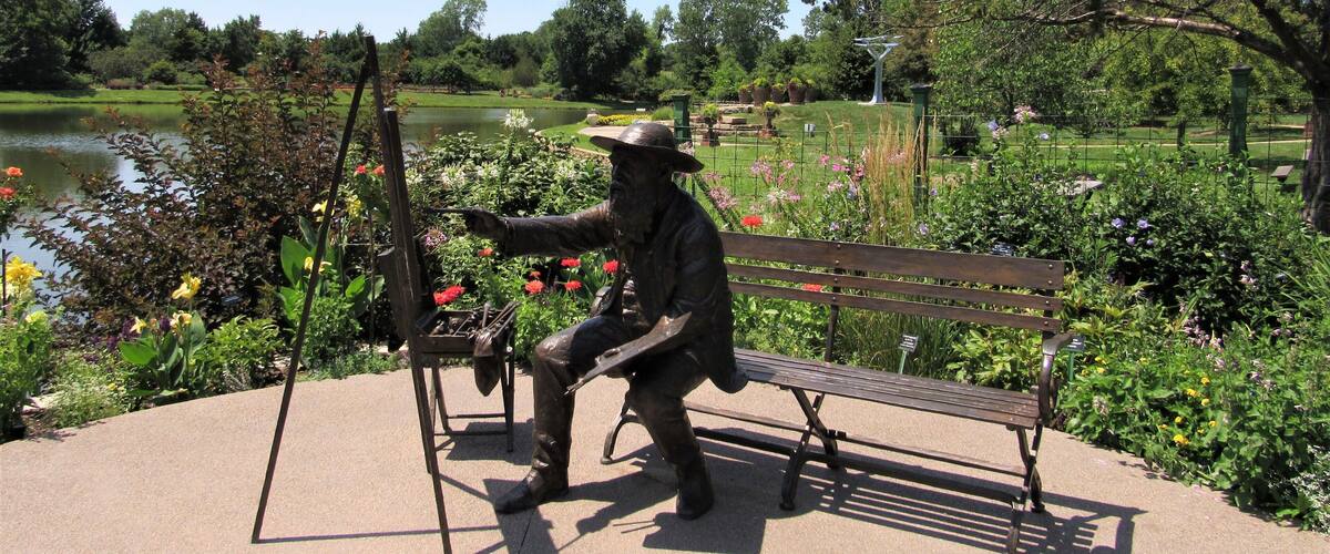 A statue of a man painting on a canvas, a bench, and a pond in a park.