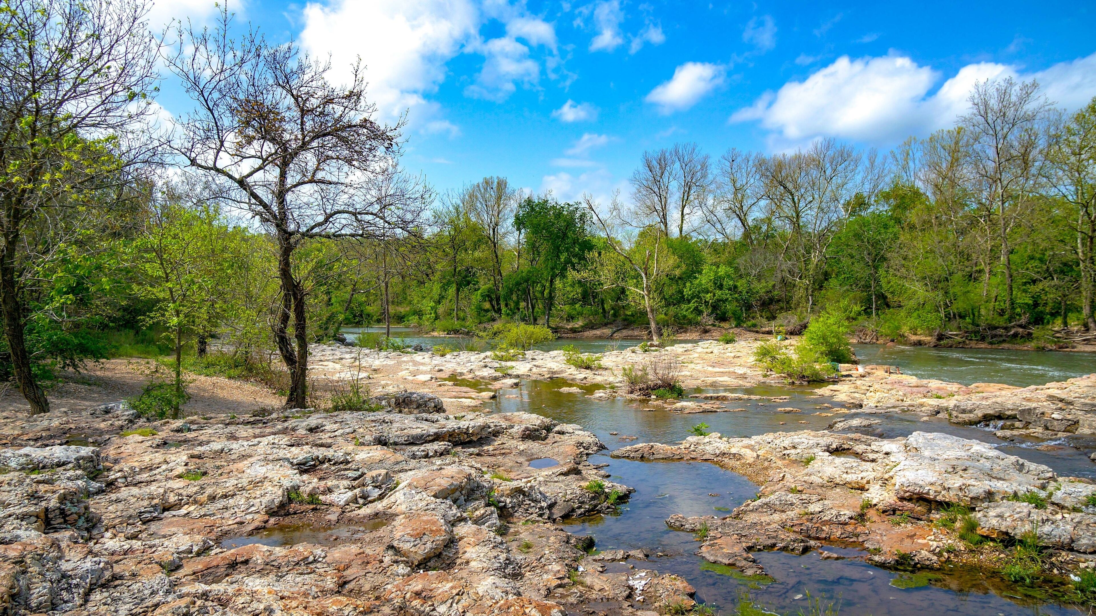 A rocky riverbed with clear water, surrounded by trees and greenery.