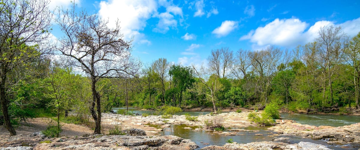 A rocky riverbed with clear water, surrounded by trees and greenery.