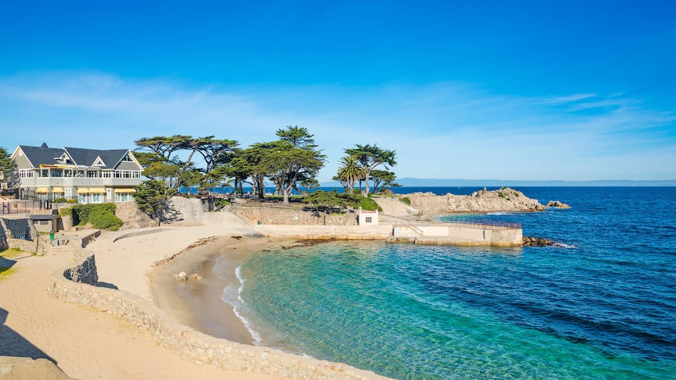 A coastal landscape with a sandy beach, clear blue waters, and a large building with a balcony overlooking the sea.