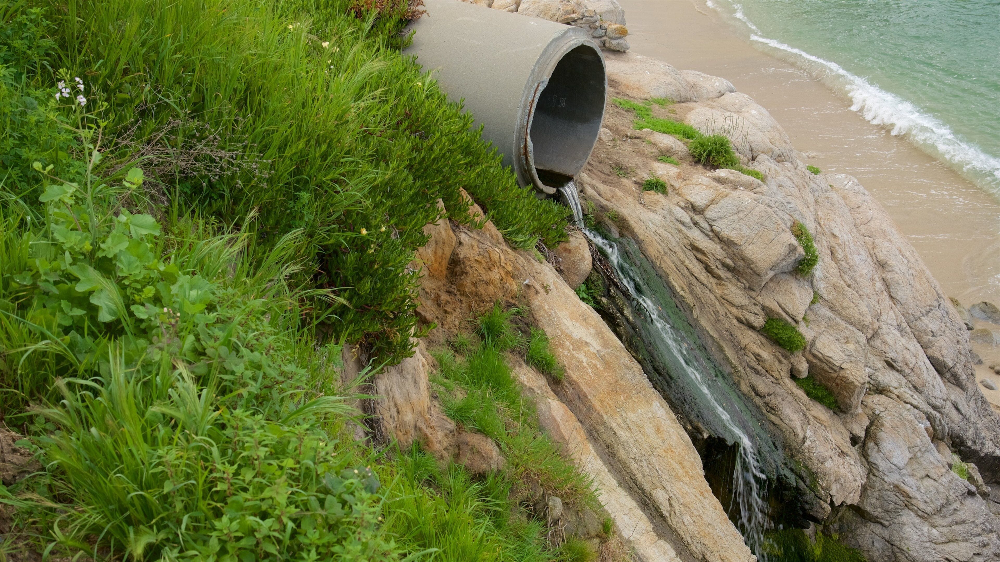 A concrete pipe discharging water into a rocky stream near a beach.