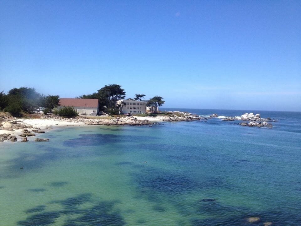 A coastal landscape with clear turquoise waters, rocky shorelines, and a building with a red roof.