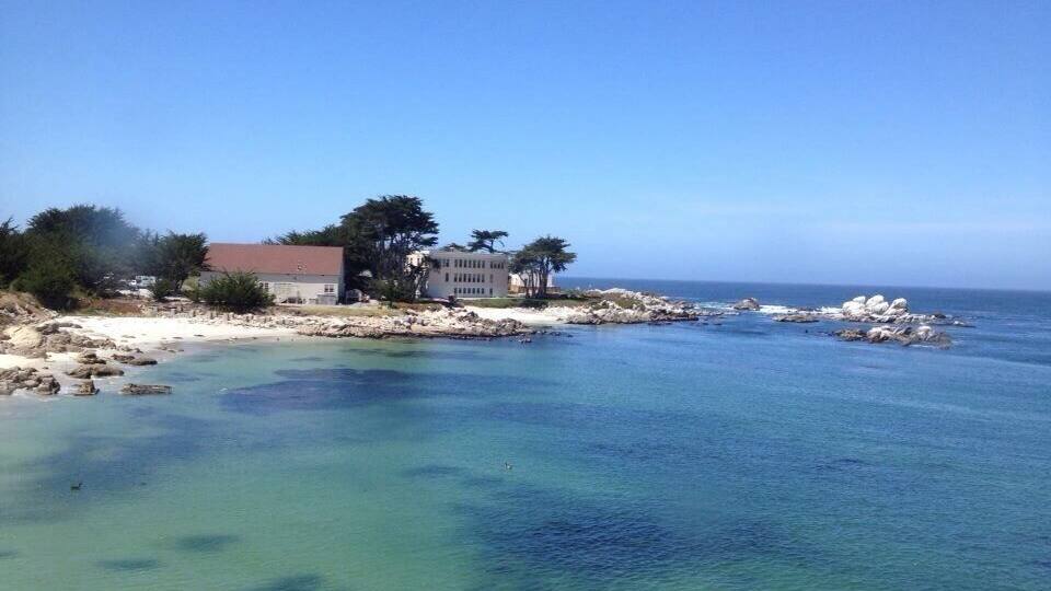 A coastal landscape with clear turquoise waters, rocky shorelines, and a building with a red roof.
