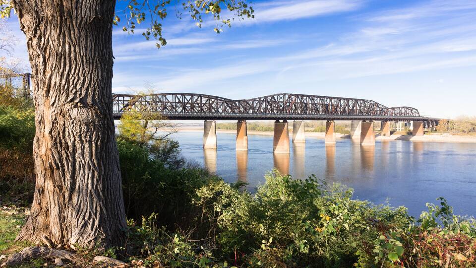 A steel bridge over a calm river, surrounded by trees and greenery.