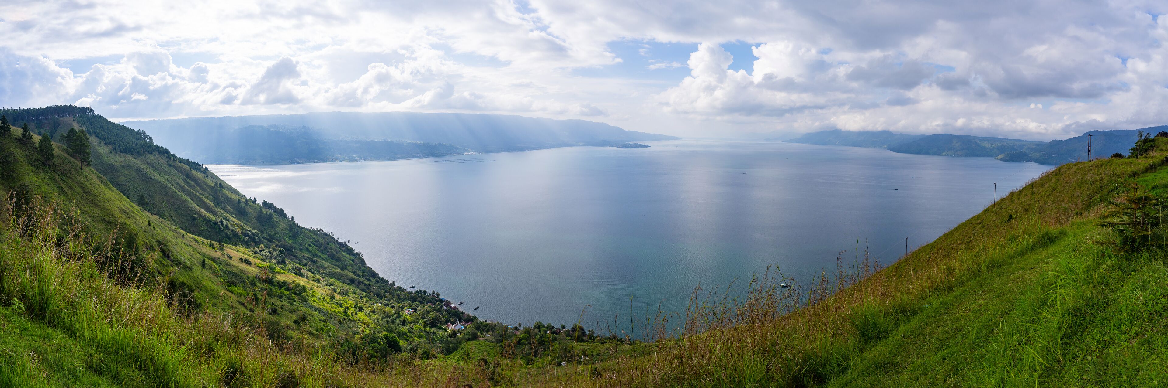 A calm lake surrounded by green hills and mountains under a partly cloudy sky.