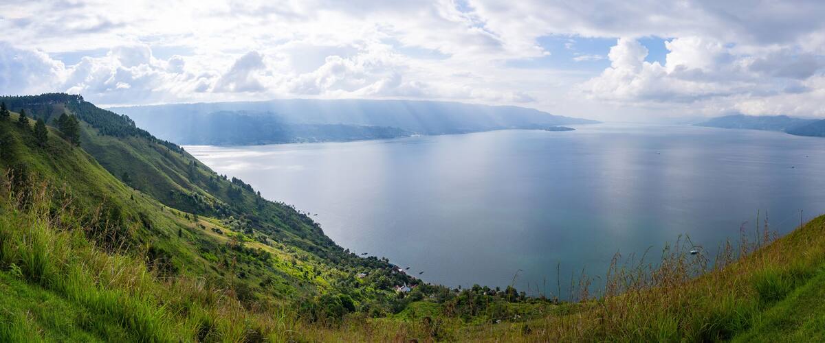 A calm lake surrounded by green hills and mountains under a partly cloudy sky.
