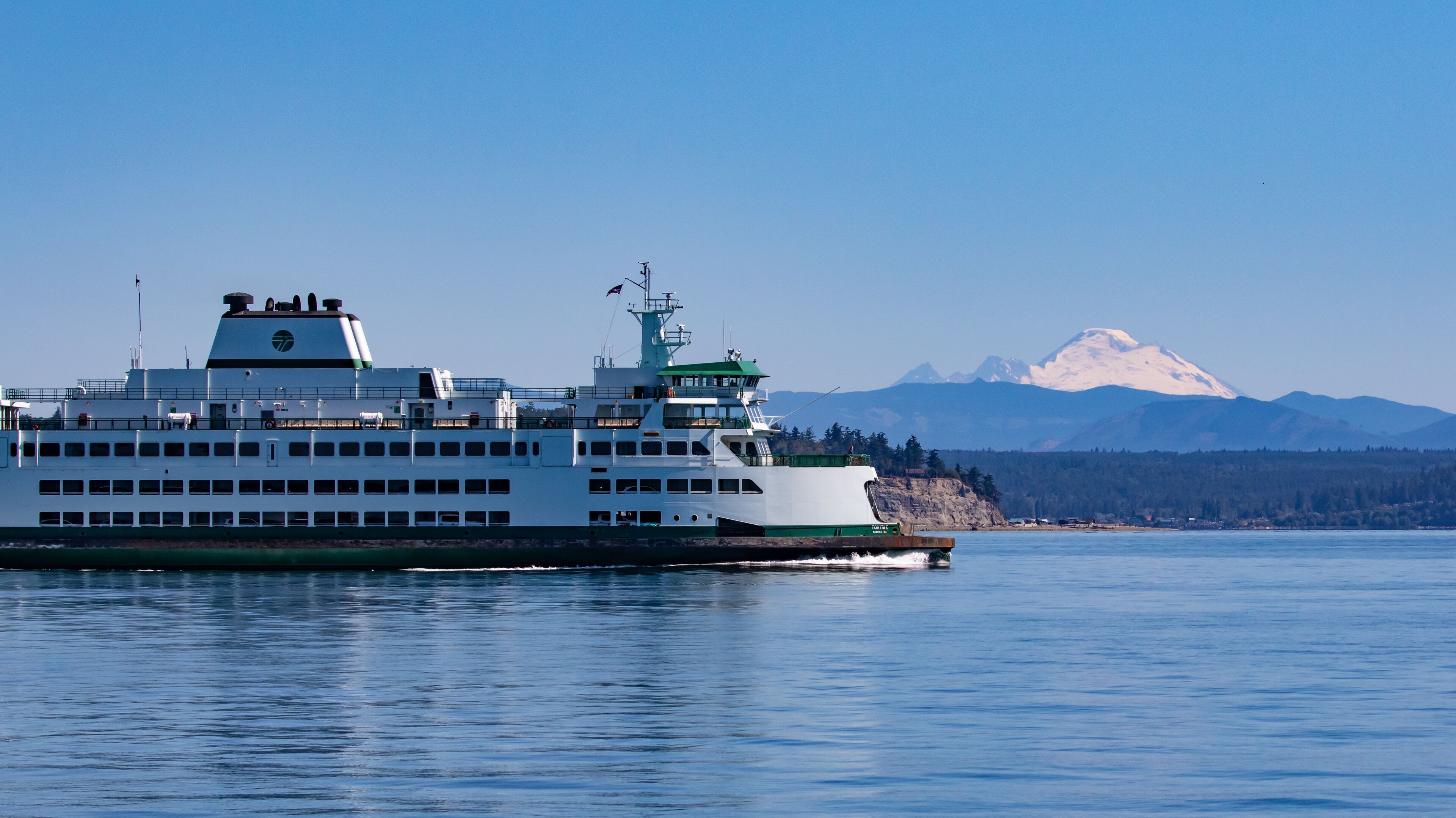 A large ferry with a green and white color scheme, docked at a port with a backdrop of snow-capped mountains and a clear blue sky.