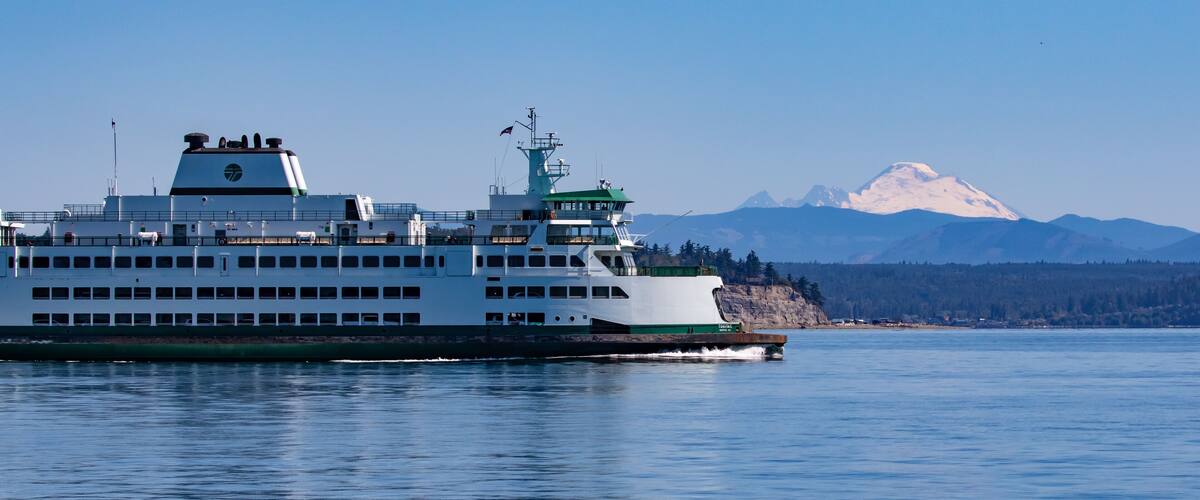 A large ferry with a green and white color scheme, docked at a port with a backdrop of snow-capped mountains and a clear blue sky.