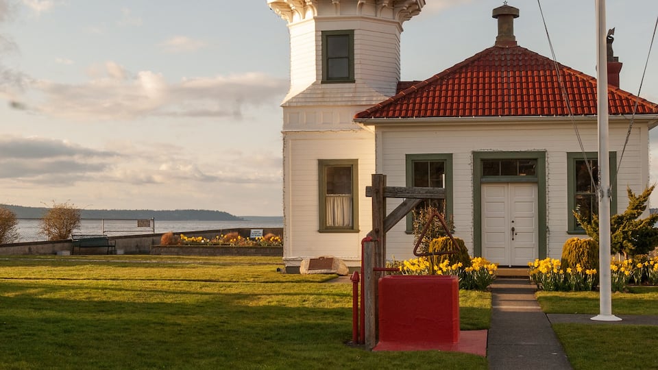 A white lighthouse with a red roof, surrounded by a well-maintained lawn and vibrant yellow flowers.