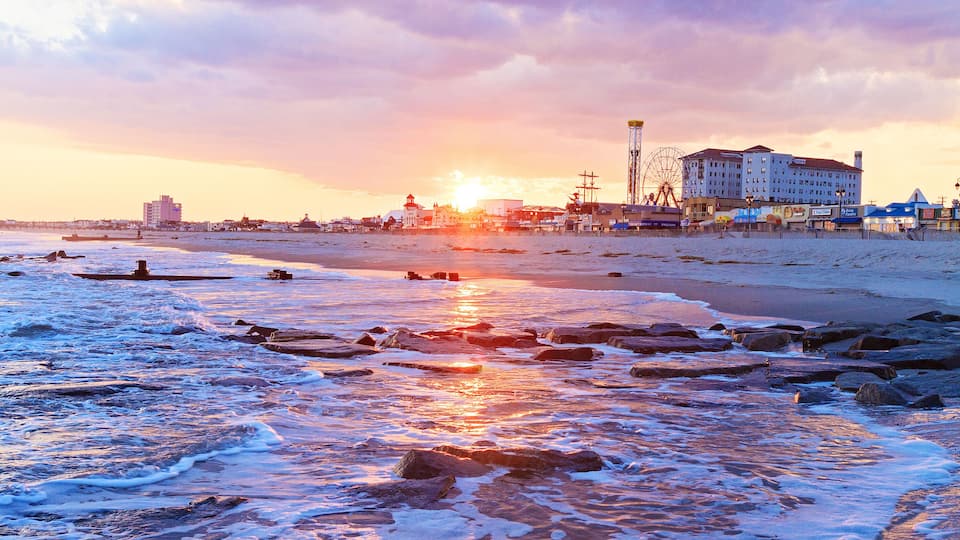 A beach with waves, a pier, and buildings in the background during sunset.