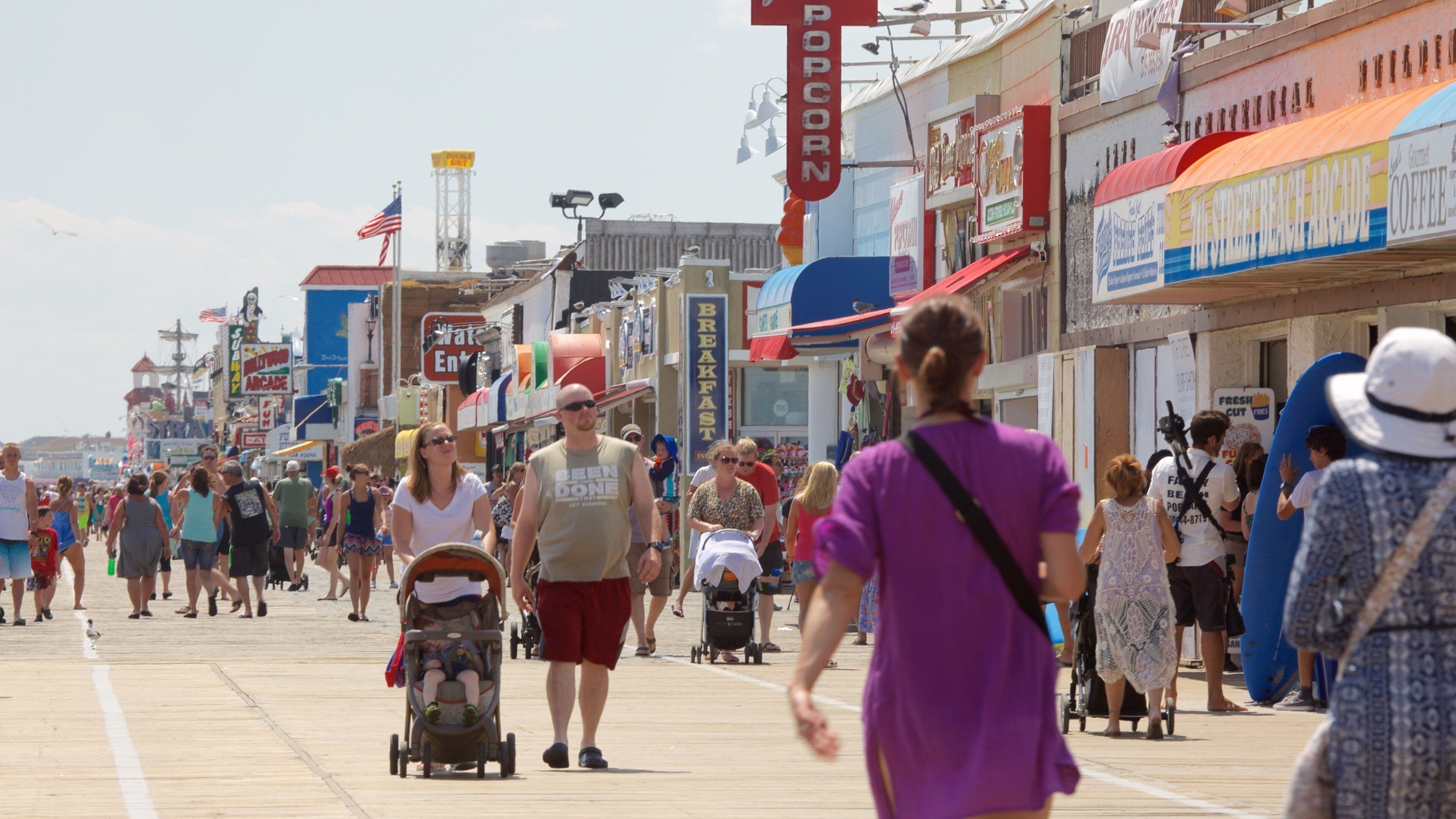 A crowded boardwalk with various shops and signs, including a popcorn stand and a coffee shop.