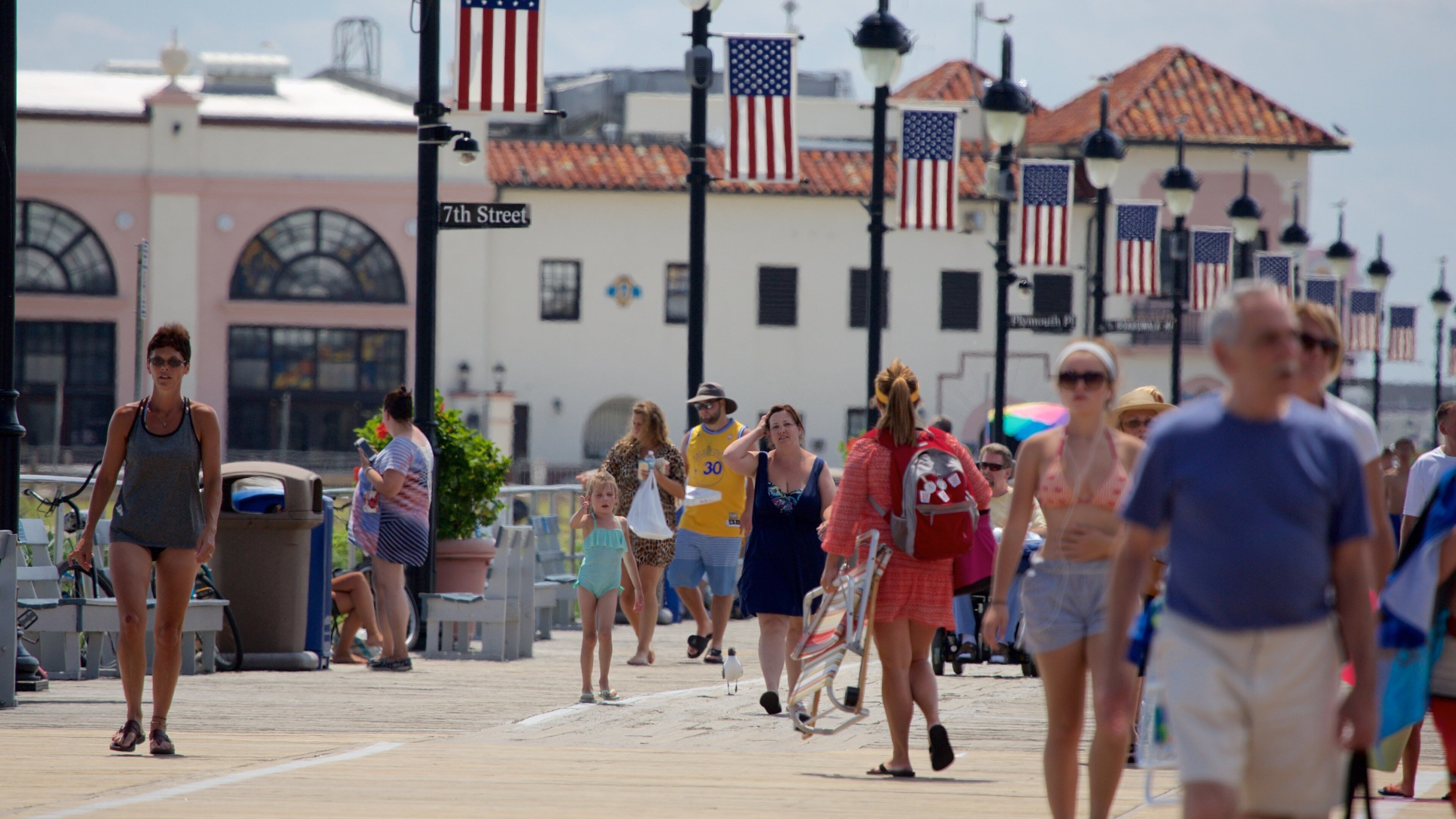 A crowded boardwalk with American flags, a sign reading "7th Street," and a building with a pink facade in the background.