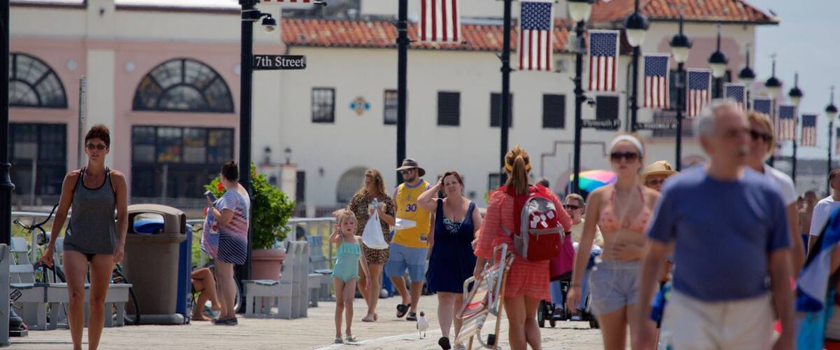 A crowded boardwalk with American flags, a sign reading "7th Street," and a building with a pink facade in the background.