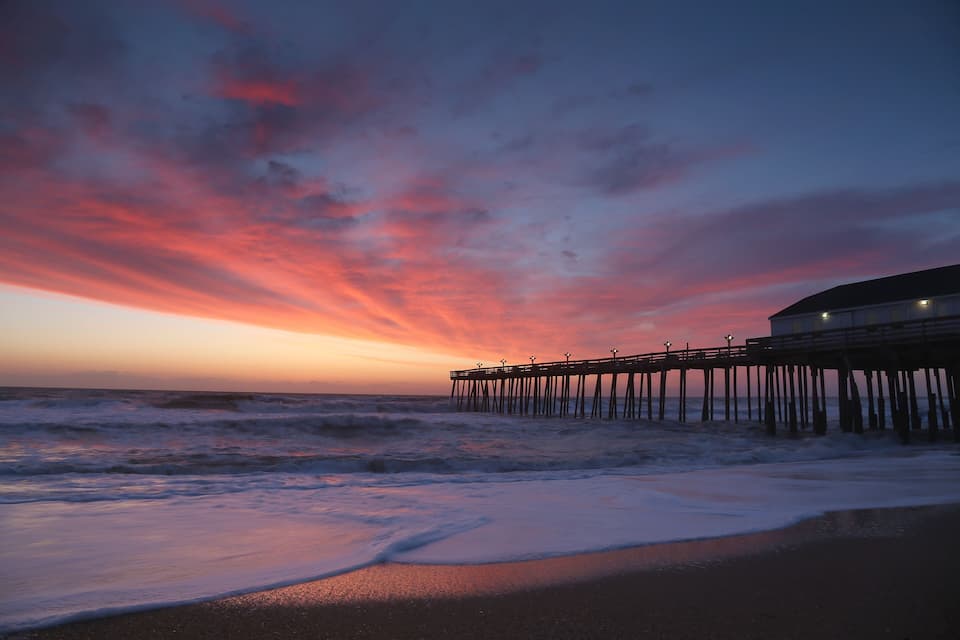 A sunset over a beach with waves, a pier, and a building.