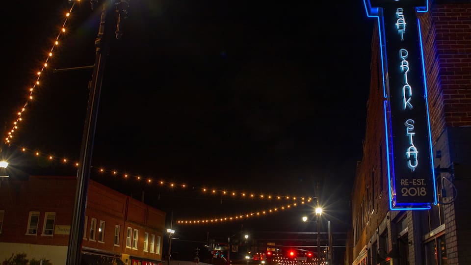 A street at night with string lights, a neon sign for 'Salem's Eat 'n Park,' and brick buildings.