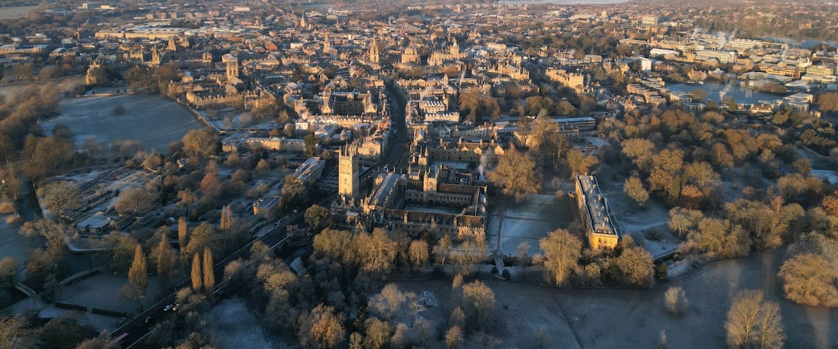 An aerial view of a historic city with dense architecture, a prominent cathedral, and surrounding green spaces.