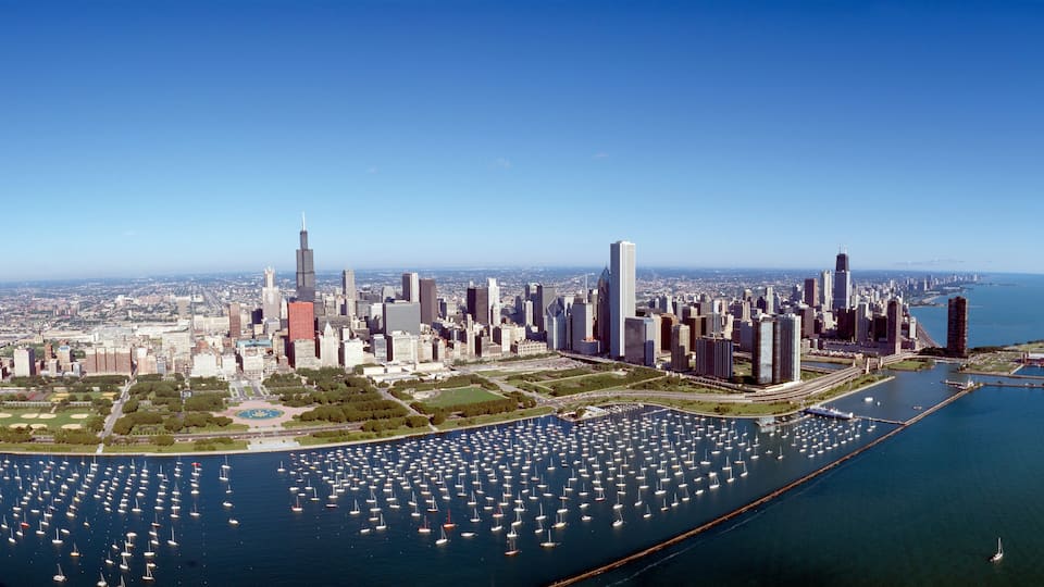 A city skyline with numerous high-rise buildings, a large body of water with numerous boats, and a clear blue sky.