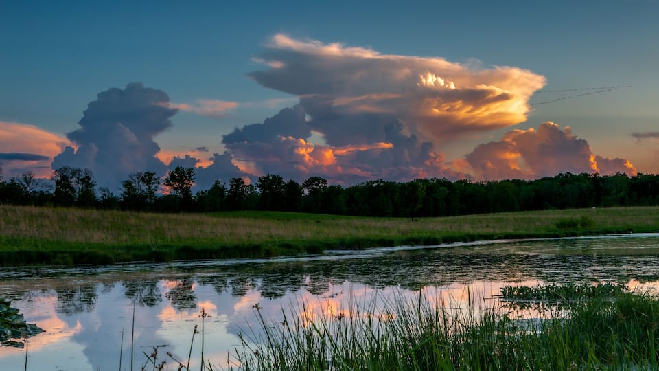 A river reflecting the sky, surrounded by grassy fields and trees.