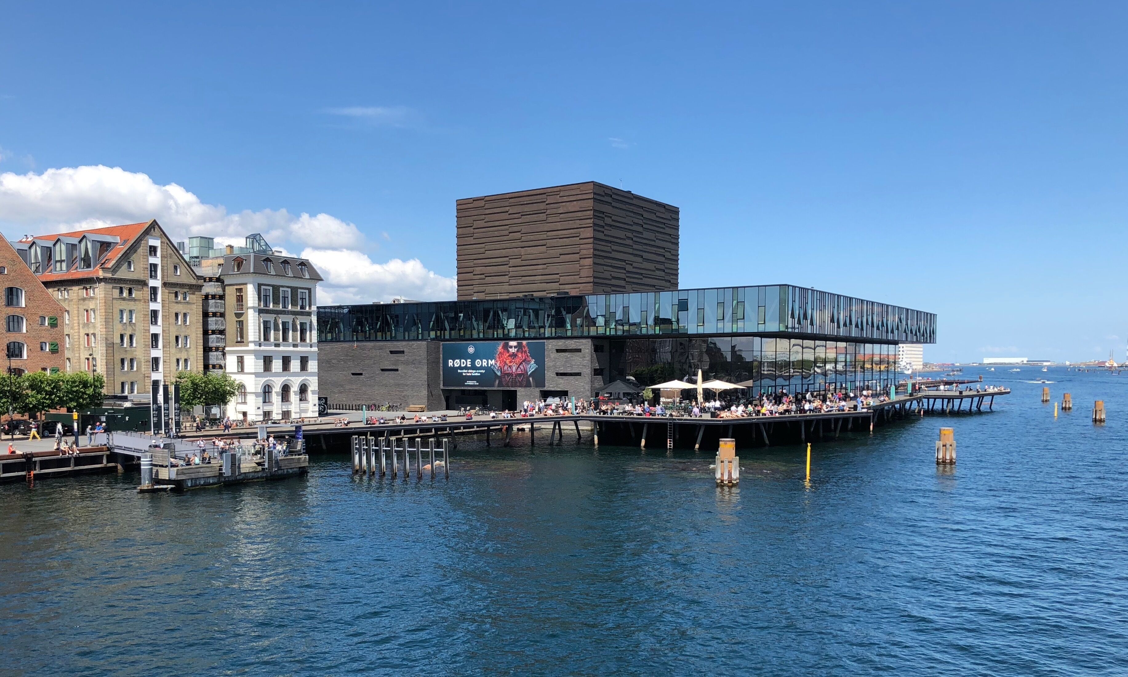 Un paseo marítimo con arquitectura moderna, que incluye un edificio de cristal y otro de ladrillo tradicional.