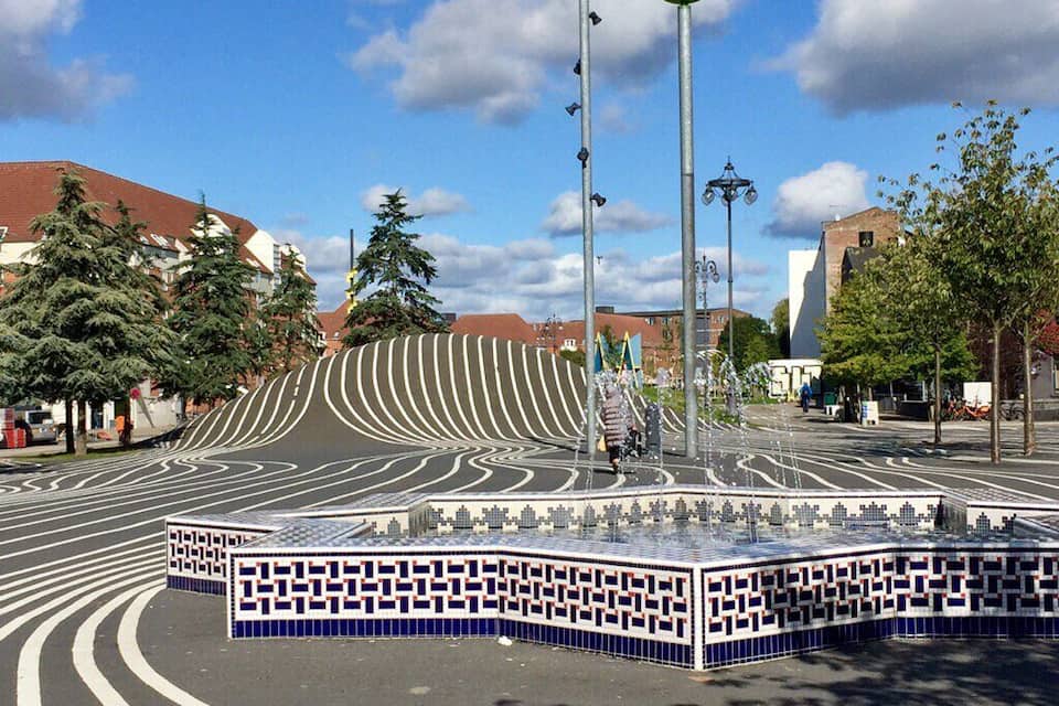 Una plaza pública con suelo de baldosas estampadas, una torre del reloj y edificios circundantes.