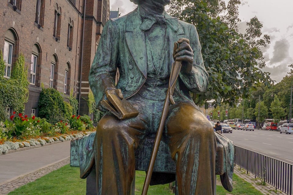 Una estàtua de bronze d'un home assegut amb un bastó, portant un barret i un abric, situada sobre un pedestal a una plaça pública amb edificis i arbres al fons.