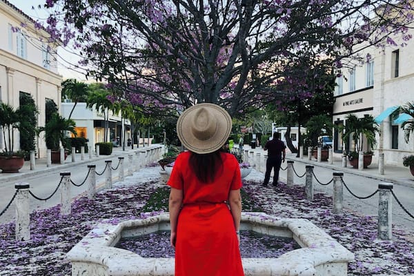 Una mujer con vestido rojo y sombrero de paja caminando hacia una fuente rodeada de flores moradas.