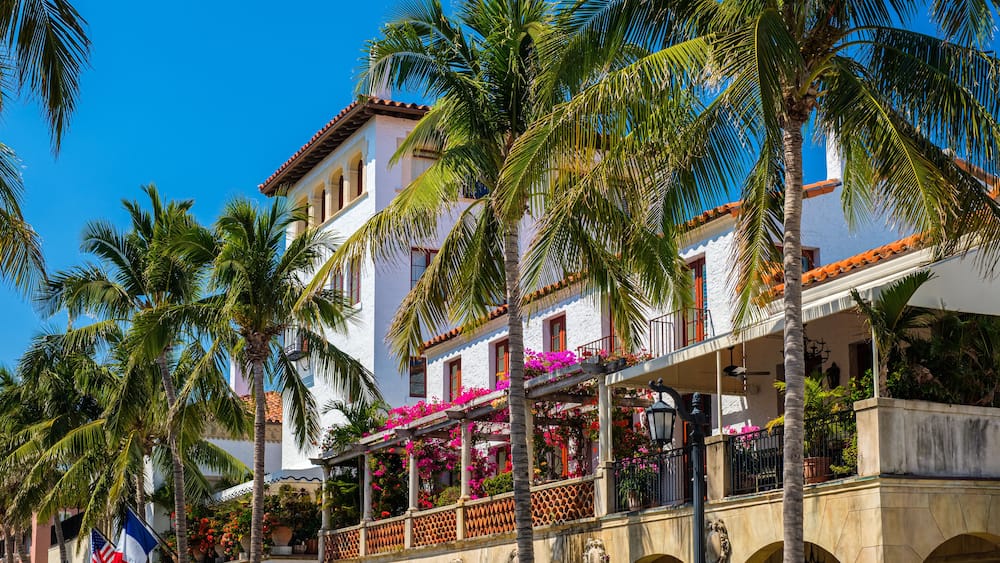 A white building with red-tiled roofs, palm trees, and balconies with pink flowers.