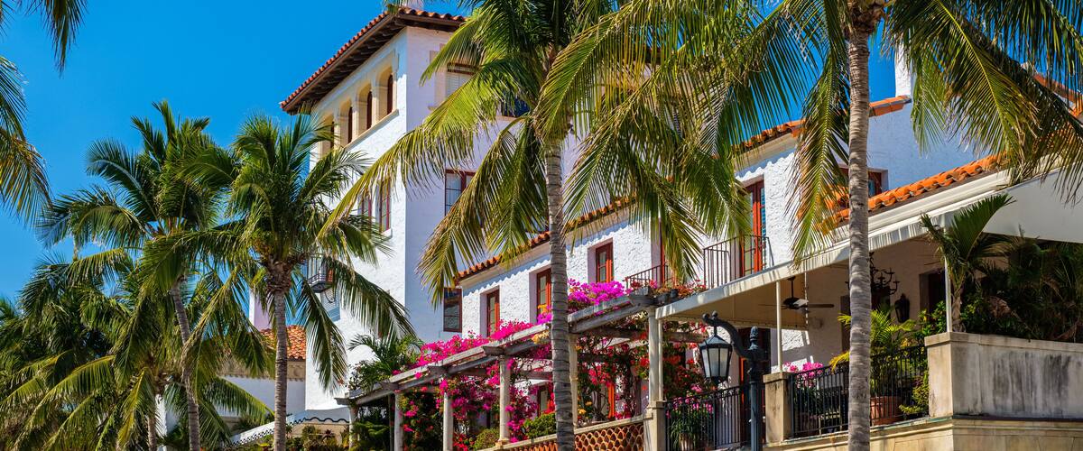 A white building with red-tiled roofs, palm trees, and balconies with pink flowers.