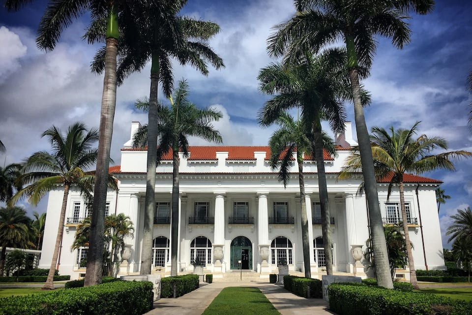 A large white building with a red roof, surrounded by palm trees and a well-maintained lawn.