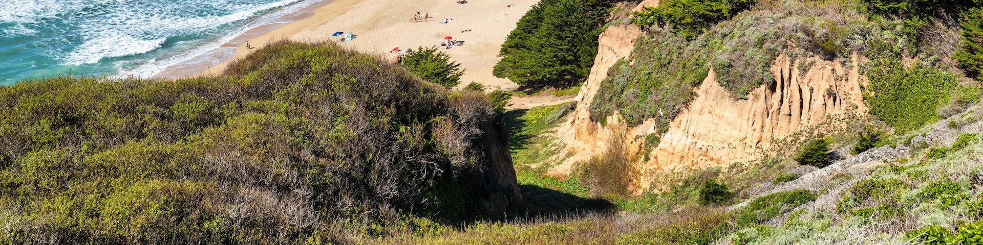 A coastal landscape with a sandy beach, rocky cliffs, and dense green vegetation.