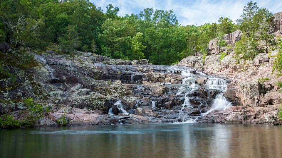A waterfall cascading over rocky terrain into a calm river, surrounded by dense green forest.