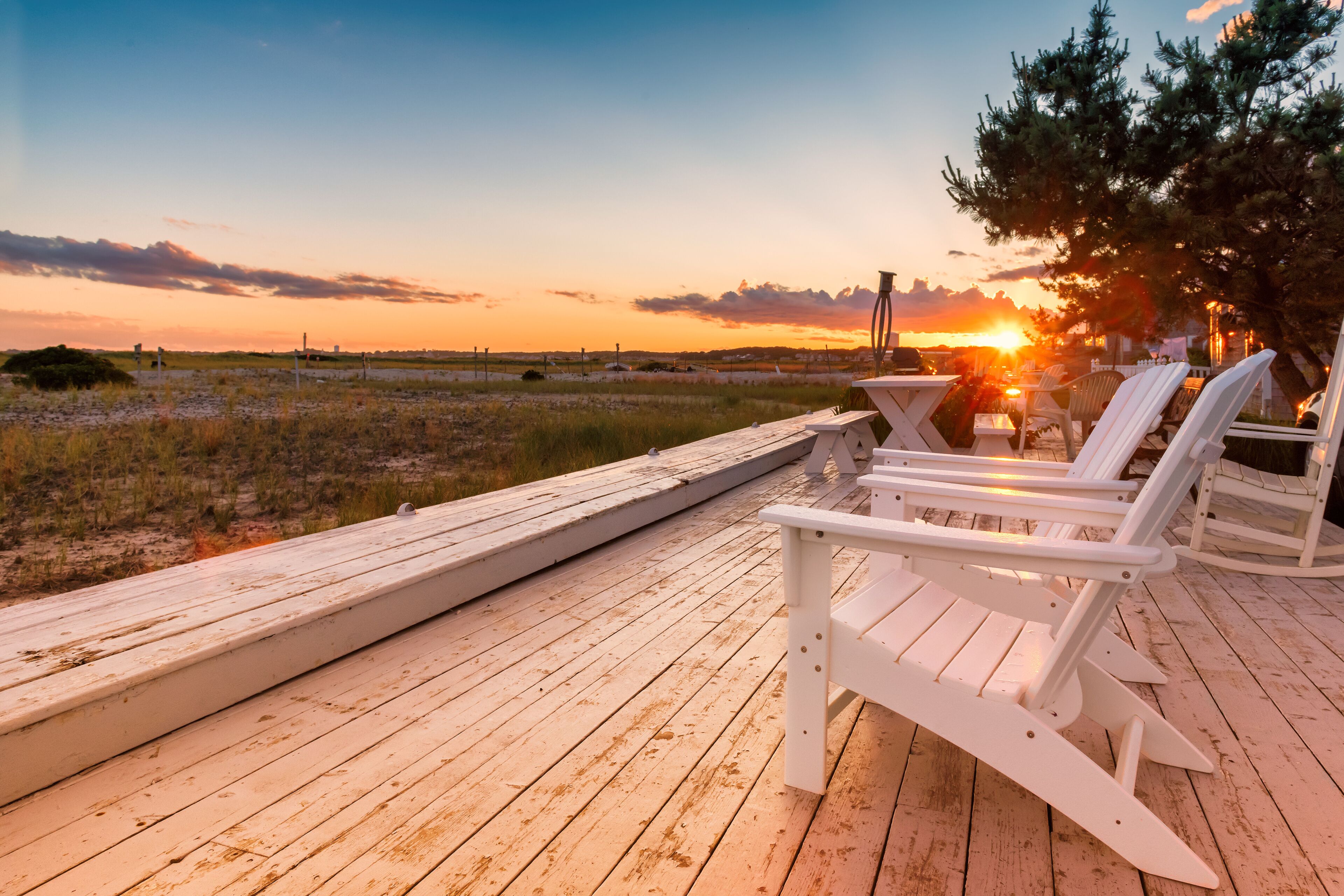 Terraza de madera con sillas Adirondack blancas frente a una playa al atardecer.