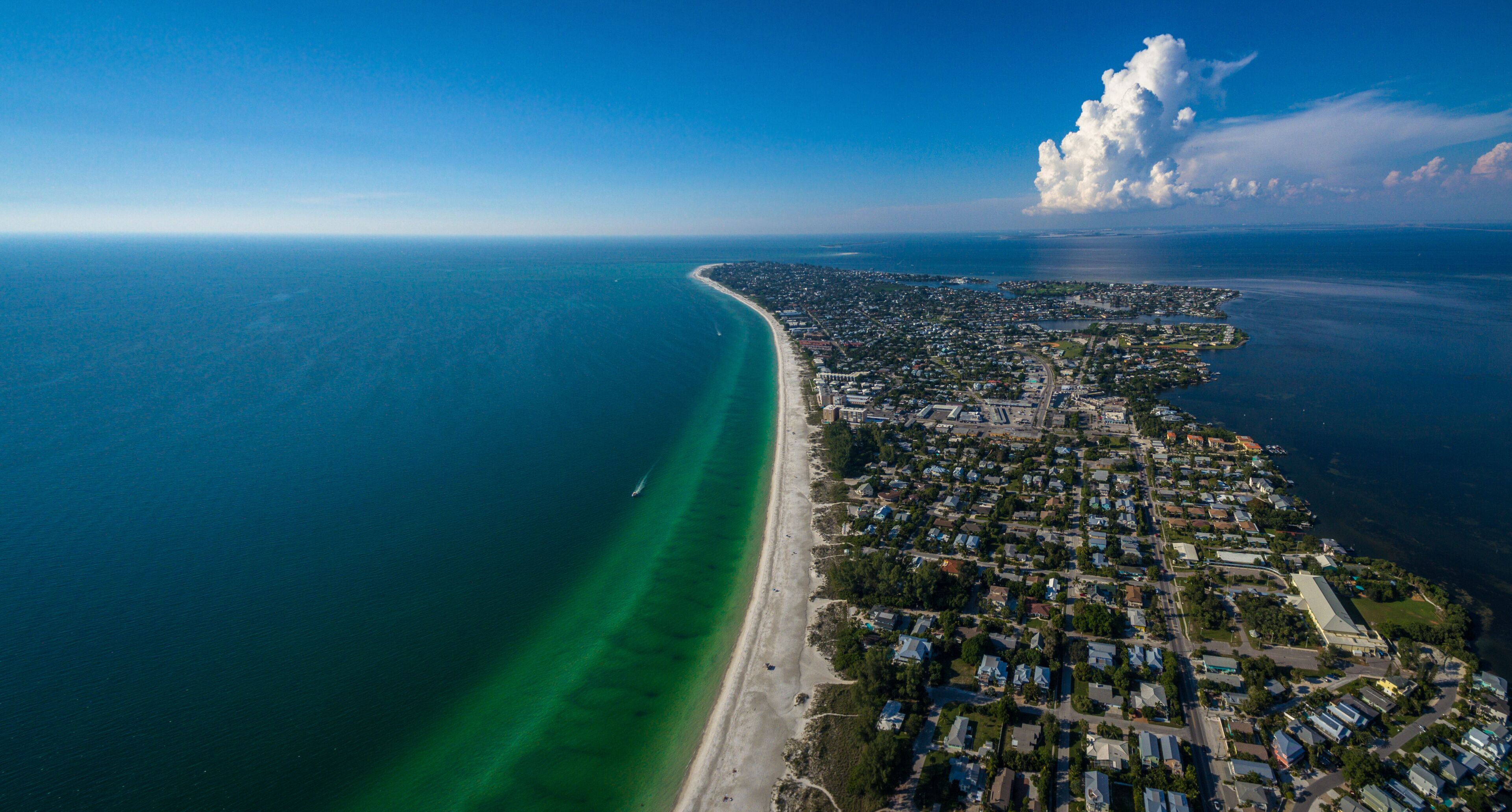 An aerial view of a coastal city with a long beachfront, dense residential area, and a large cloud formation.