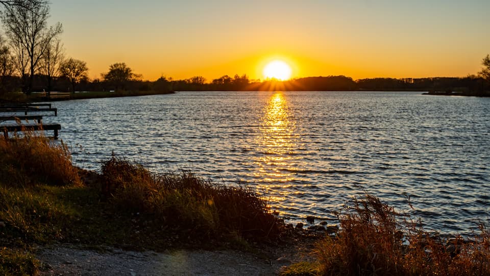 A sunset over a calm lake with a bench and grassy banks.