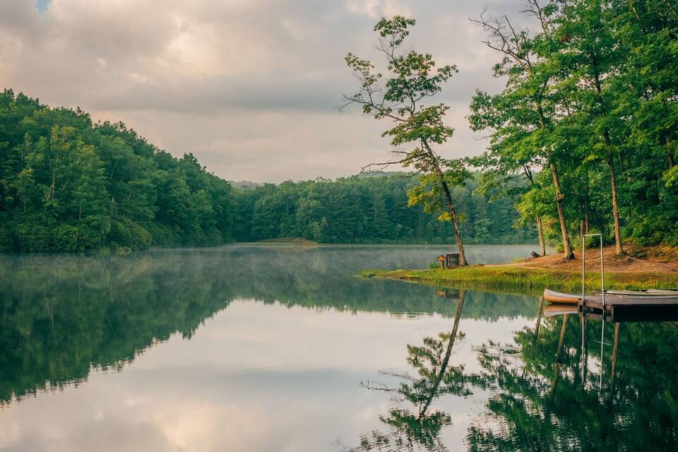 A calm lake with reflections of trees and clouds, surrounded by dense forest and a dock with a boat.