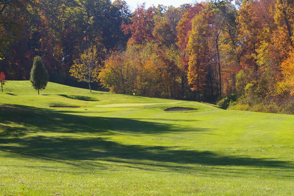 A golf course with a green fairway, sand bunkers, and trees with autumn foliage.