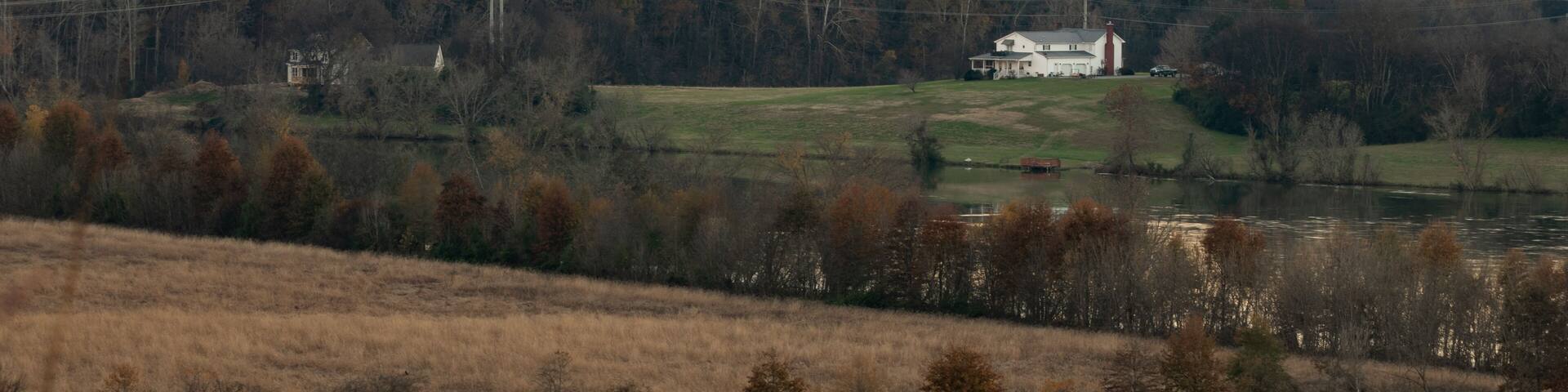 A rural landscape with rolling hills, a river, and a white house surrounded by trees.