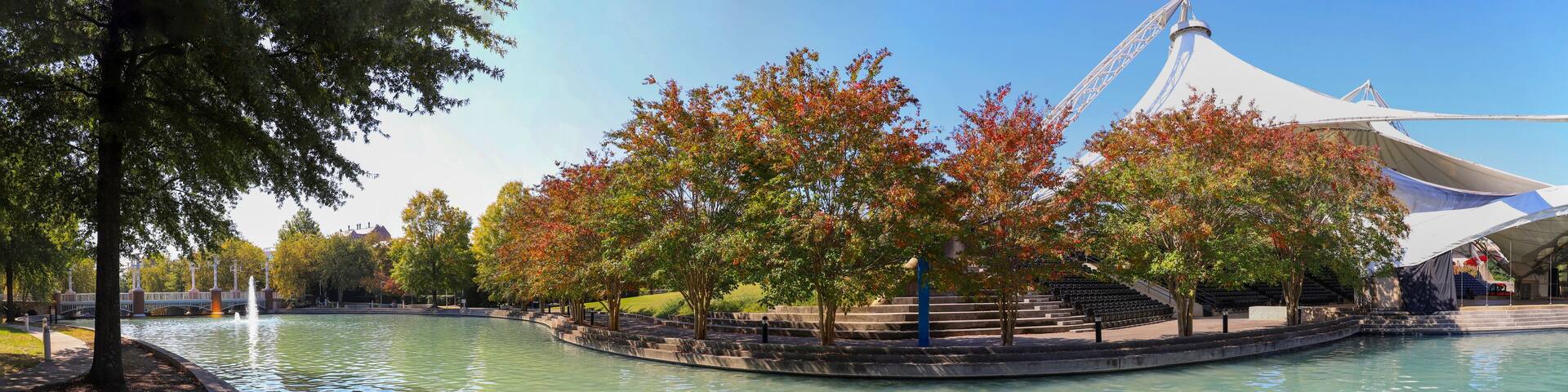 A park with a reflective water body, trees with autumn foliage, and a white tent-like structure.