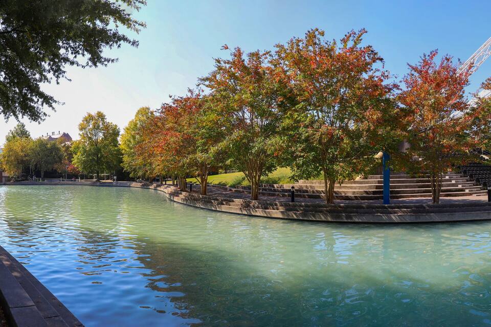 A park with a reflective water body, trees with autumn foliage, and a white tent-like structure.