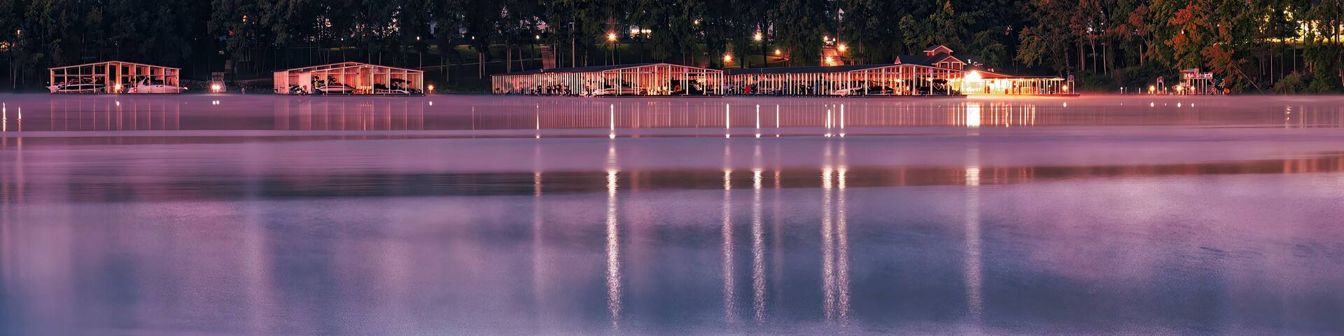 Un lac paisible au crépuscule, avec des quais et des bâtiments illuminés qui se reflètent dans l’eau.
